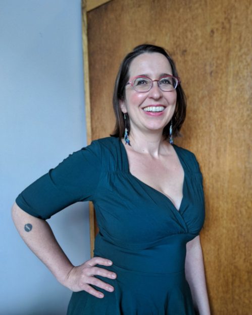 Mazarine Treyz a pale person with glasses smiling to the right with a wooden background